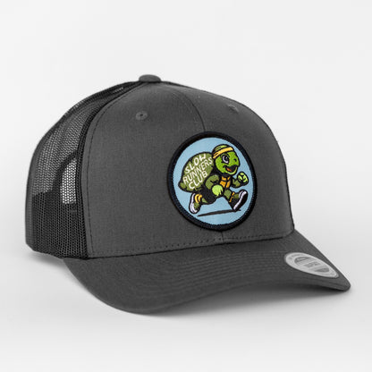 Angled to one side, the charcoal Slow Runners Club trucker hat pairs black mesh with its running turtle patch.