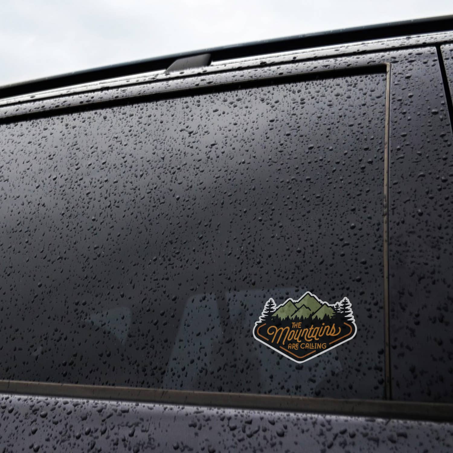 Rain beads cover a dark car window where The Mountains Are Calling sticker sits low on the glass.