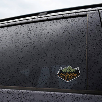 Rain beads cover a dark car window where The Mountains Are Calling sticker sits low on the glass.