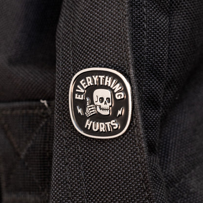 Clipped to a dark backpack strap, the Everything Hurts enamel pin shows its skull facing forward between stitched seams.