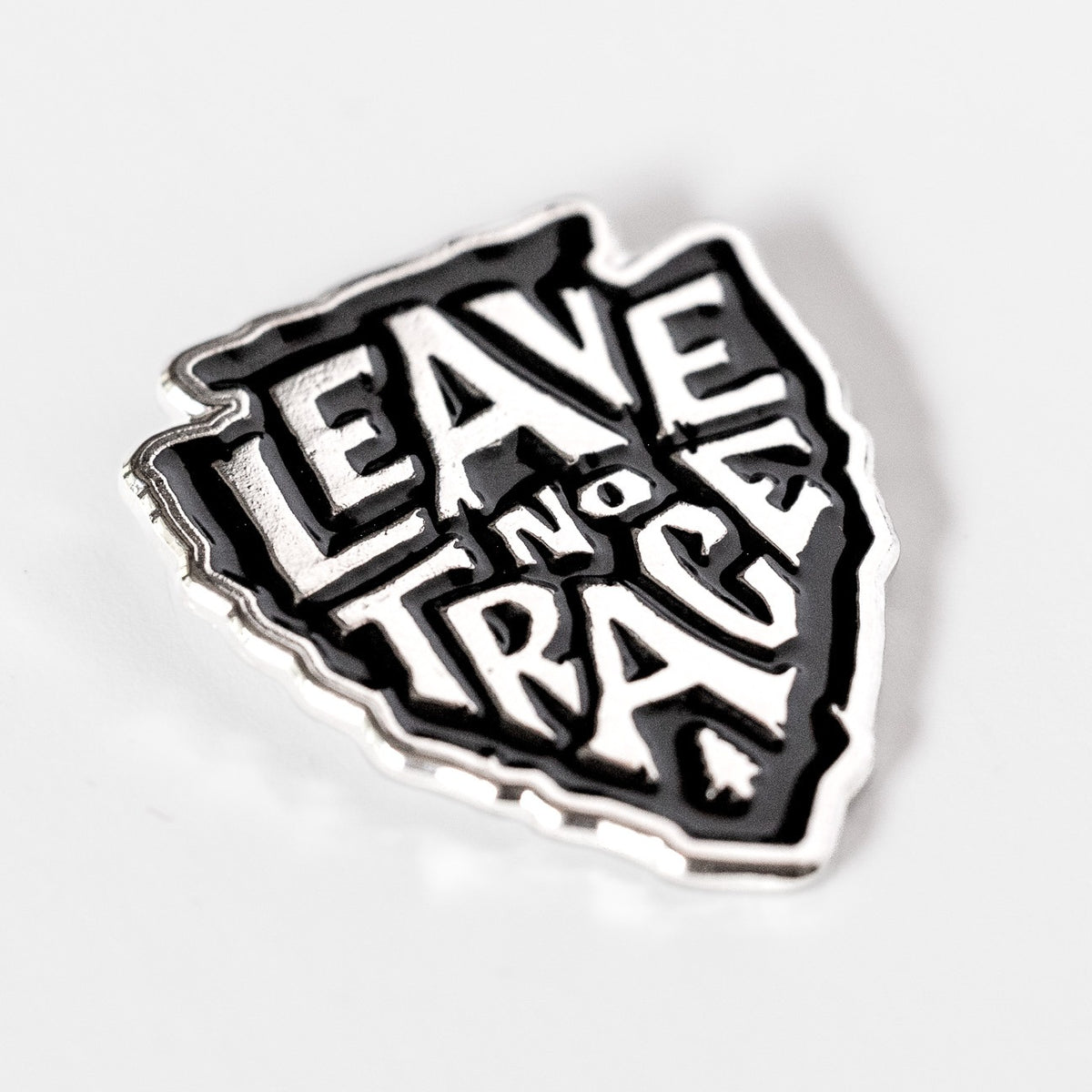 Tilted on a plain white background, the arrowhead shaped Leave No Trace pin shows its black and silver lettering.