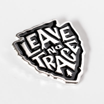 Tilted on a plain white background, the arrowhead shaped Leave No Trace pin shows its black and silver lettering.