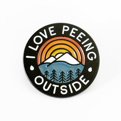 Round and black, the I Love Peeing Outside Pin displays a sunset mountain scene above pine trees and its full text.