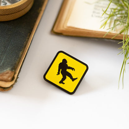 Beside a brass compass and old book pages, the yellow Bigfoot Crossing pin rests near trailing pine needles.