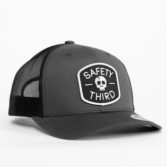 Set against a plain white background, the grey Safety Third trucker hat shows its skull patch and black mesh back.