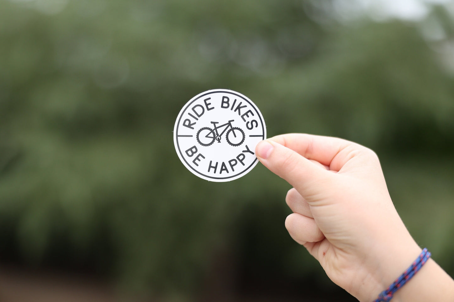 A wrist with a braided bracelet holds up the small white Ride Bikes Be Happy sticker outdoors.