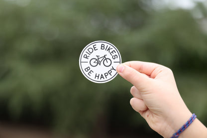 A wrist with a braided bracelet holds up the small white Ride Bikes Be Happy sticker outdoors.