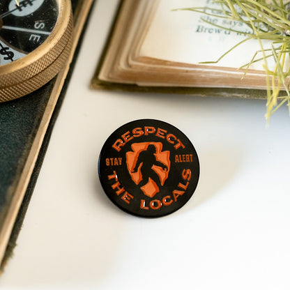 Resting on white between an open book and a brass compass, the Respect The Locals pin displays its orange Bigfoot mark.