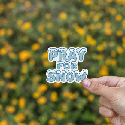 Yellow wildflowers blur behind fingers holding the Pray For Snow sticker steady.