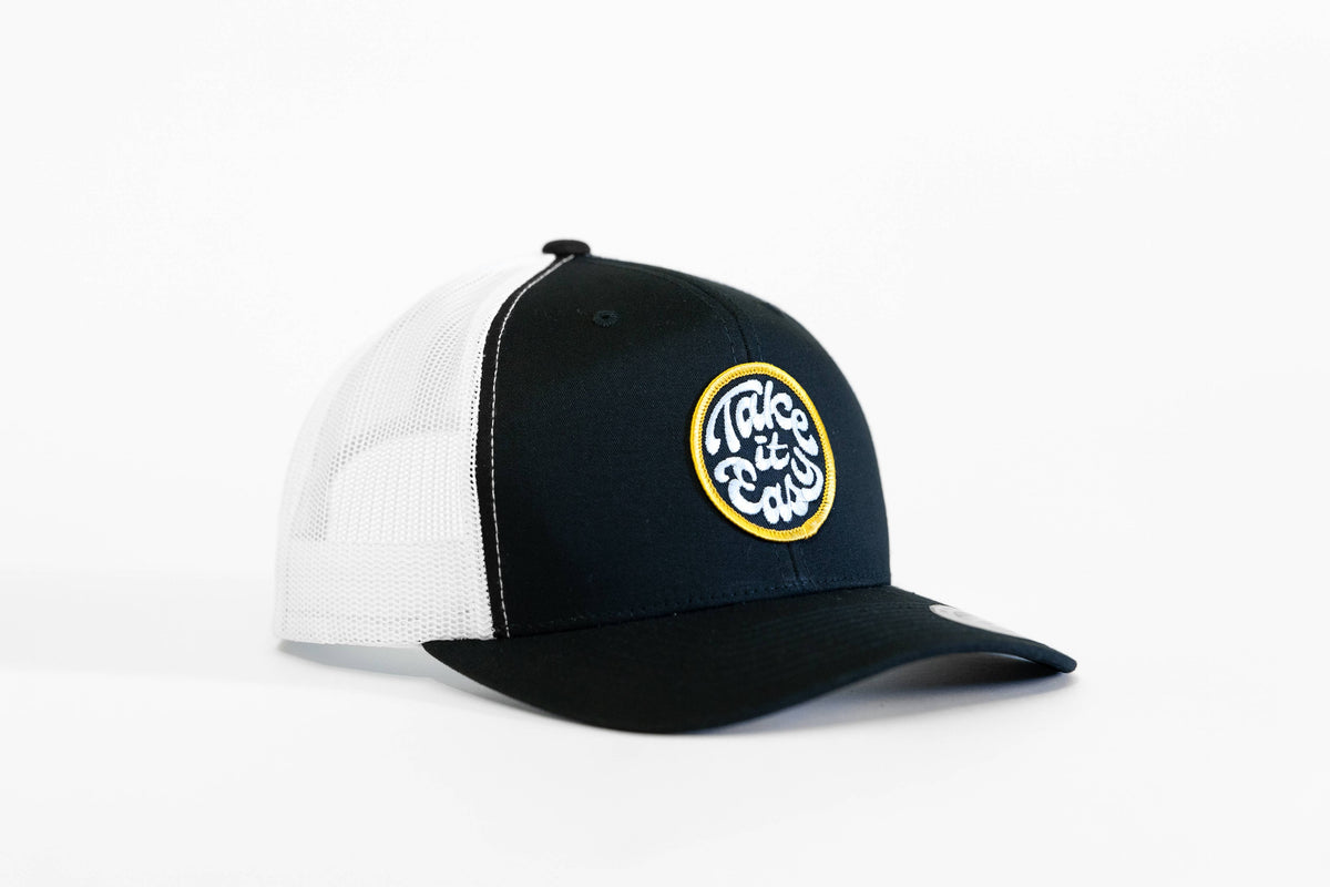 Black and white Take it Easy trucker hat angled on a plain backdrop with its round yellow patch in view.