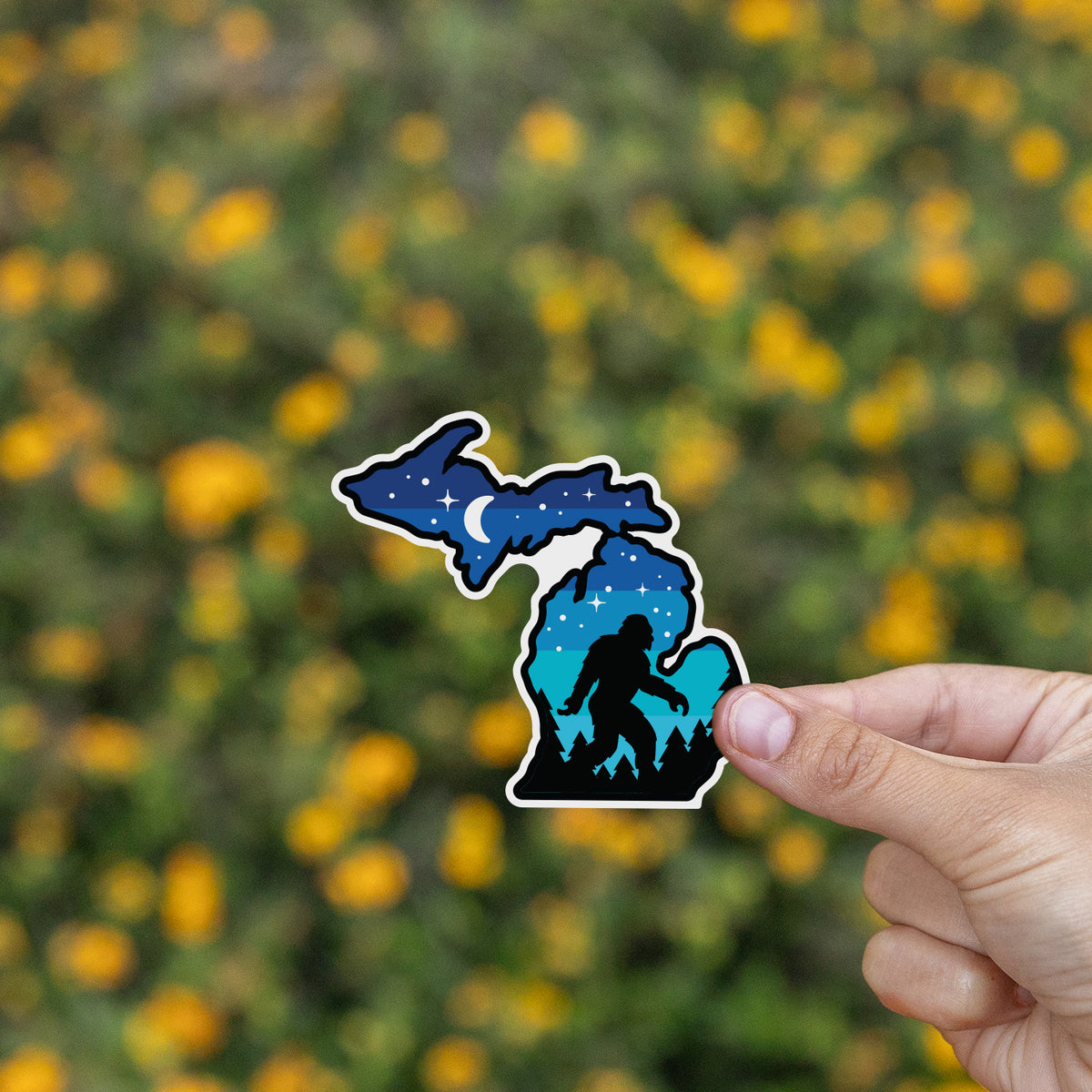 A hand lifts the Michigan Sasquatch sticker in front of a blurred field of small yellow wildflowers.