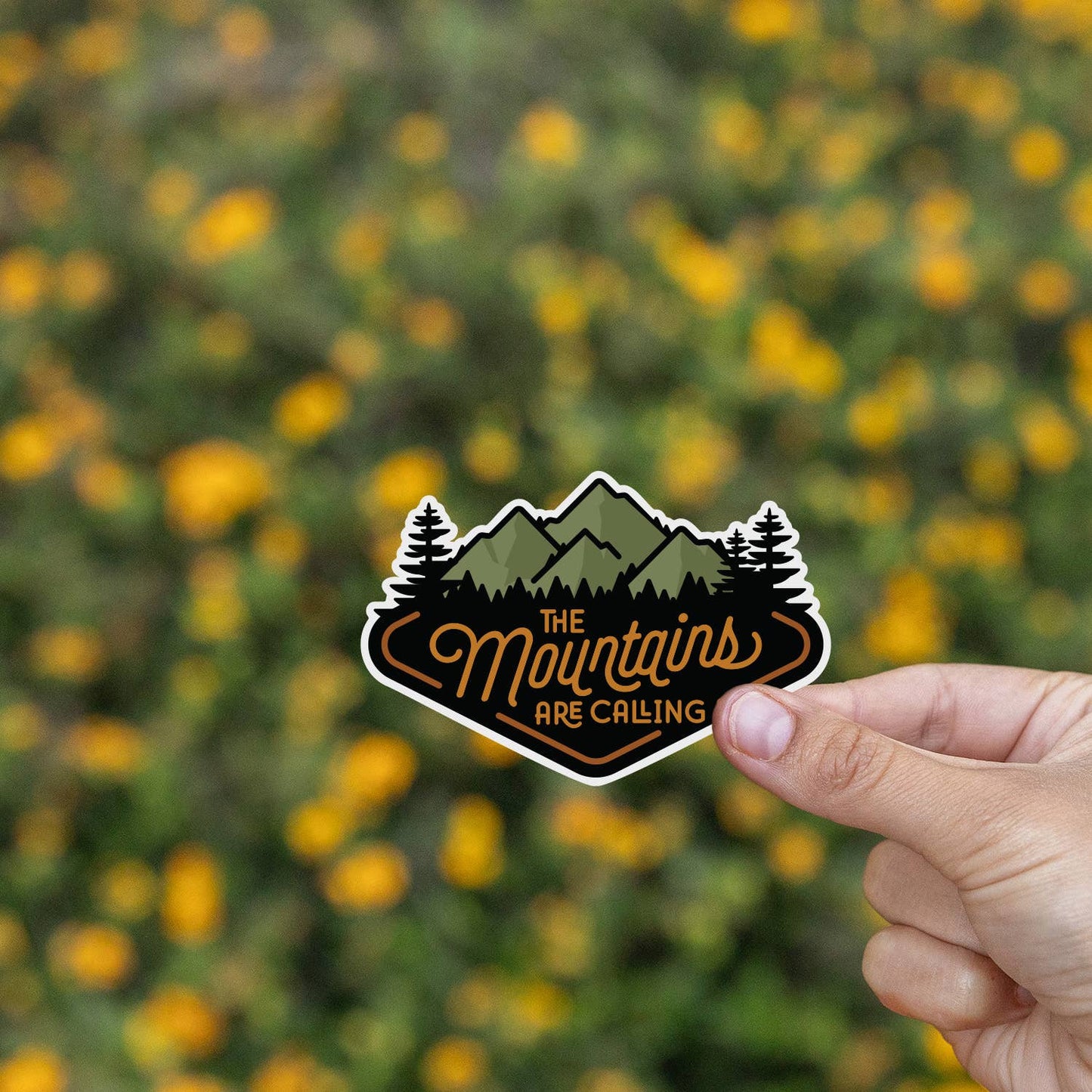 Yellow wildflowers blur behind a hand pinching The Mountains Are Calling sticker.