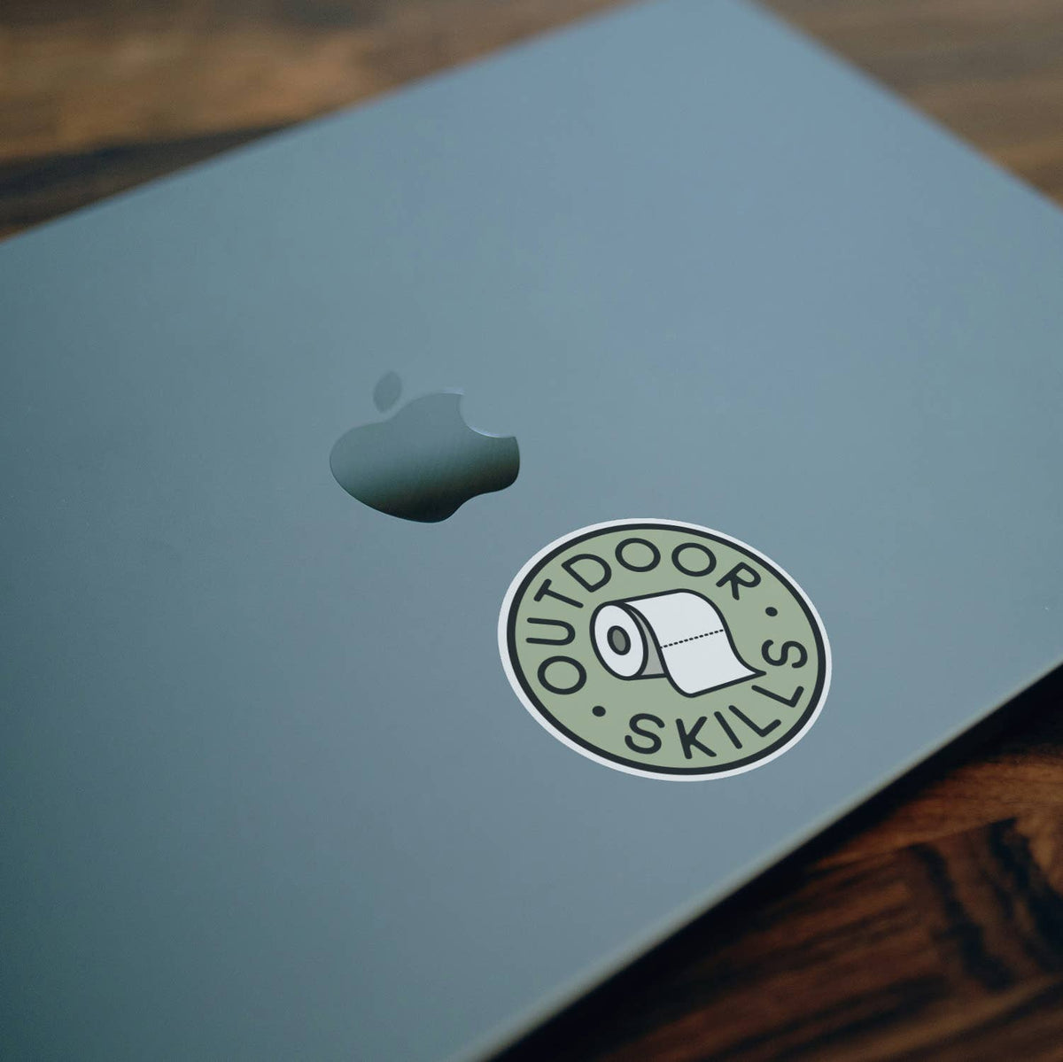 Stuck near the logo on a green laptop lid, the Outdoor Skills sticker sits on a dark wood desk.