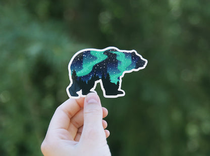 Raised high in one hand, the Northern Lights Grizzly Bear sticker glows teal against deep green bokeh.