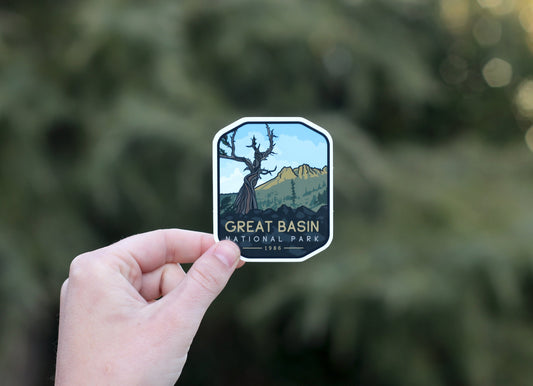 A hand holds the Great Basin National Park sticker against blurred evergreen boughs, bristlecone pine and peaks in view.
