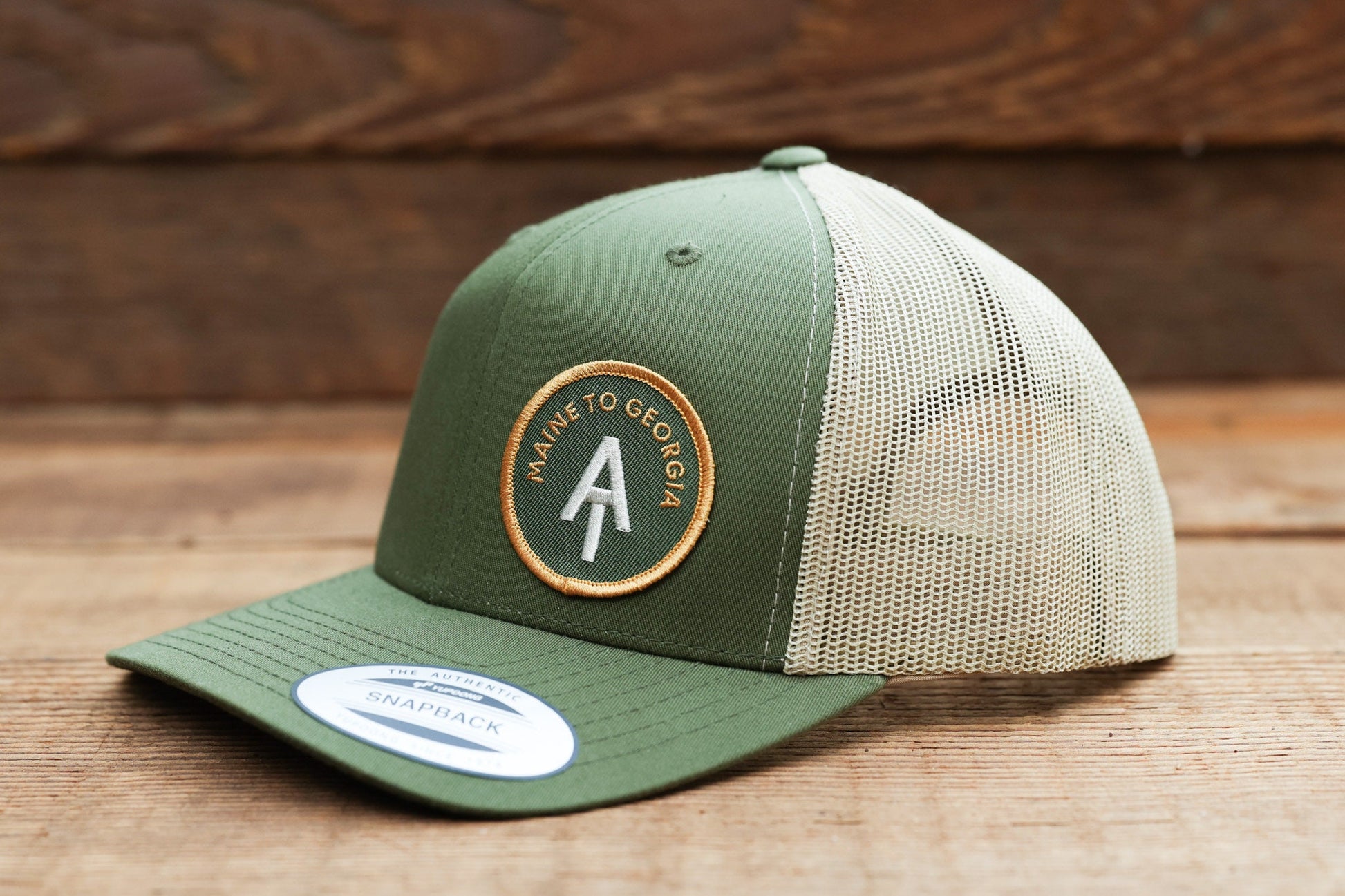 Set on wood boards, the green Appalachian Trail trucker hat tips its brim toward the left with mesh in view.