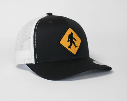 Black crown and white mesh carry the Bigfoot Crossing trucker hat and its yellow sasquatch sign patch.