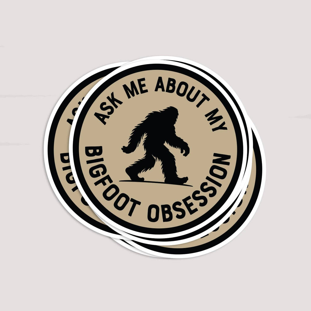 Three Ask Me About My Bigfoot Obsession stickers fan out in a loose stack on a pale surface.