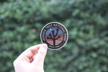 Fingers grip the Joshua Tree National Park sticker close to a dense dark hedge in flat daylight.