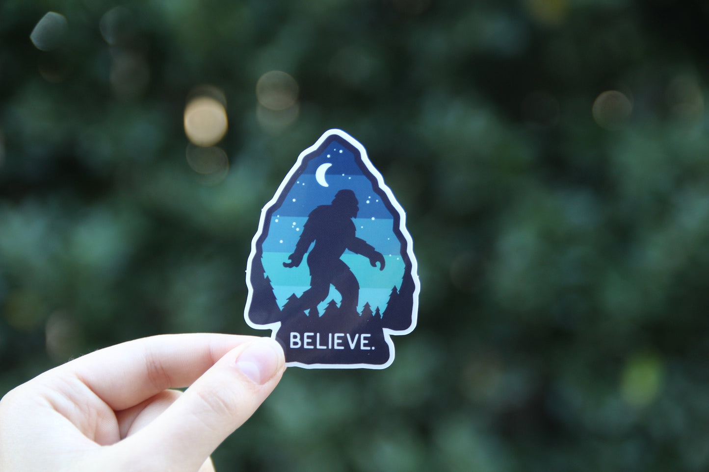 Bokeh dots glow in a dark hedge behind the Bigfoot Believe sticker raised in one hand.