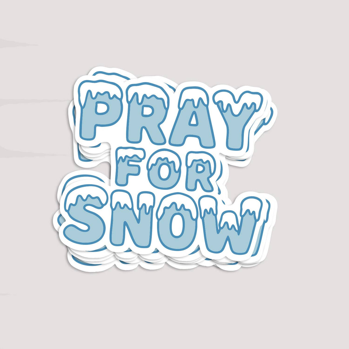 A stack of Pray For Snow stickers rests on a pale grey surface, edges layered under the top one.