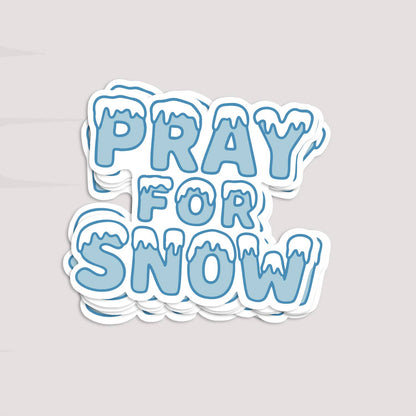 A stack of Pray For Snow stickers rests on a pale grey surface, edges layered under the top one.