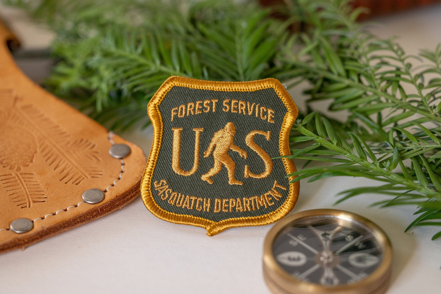 Laid on a white table, the US Sasquatch Department patch sits between stamped leather and cedar branches.