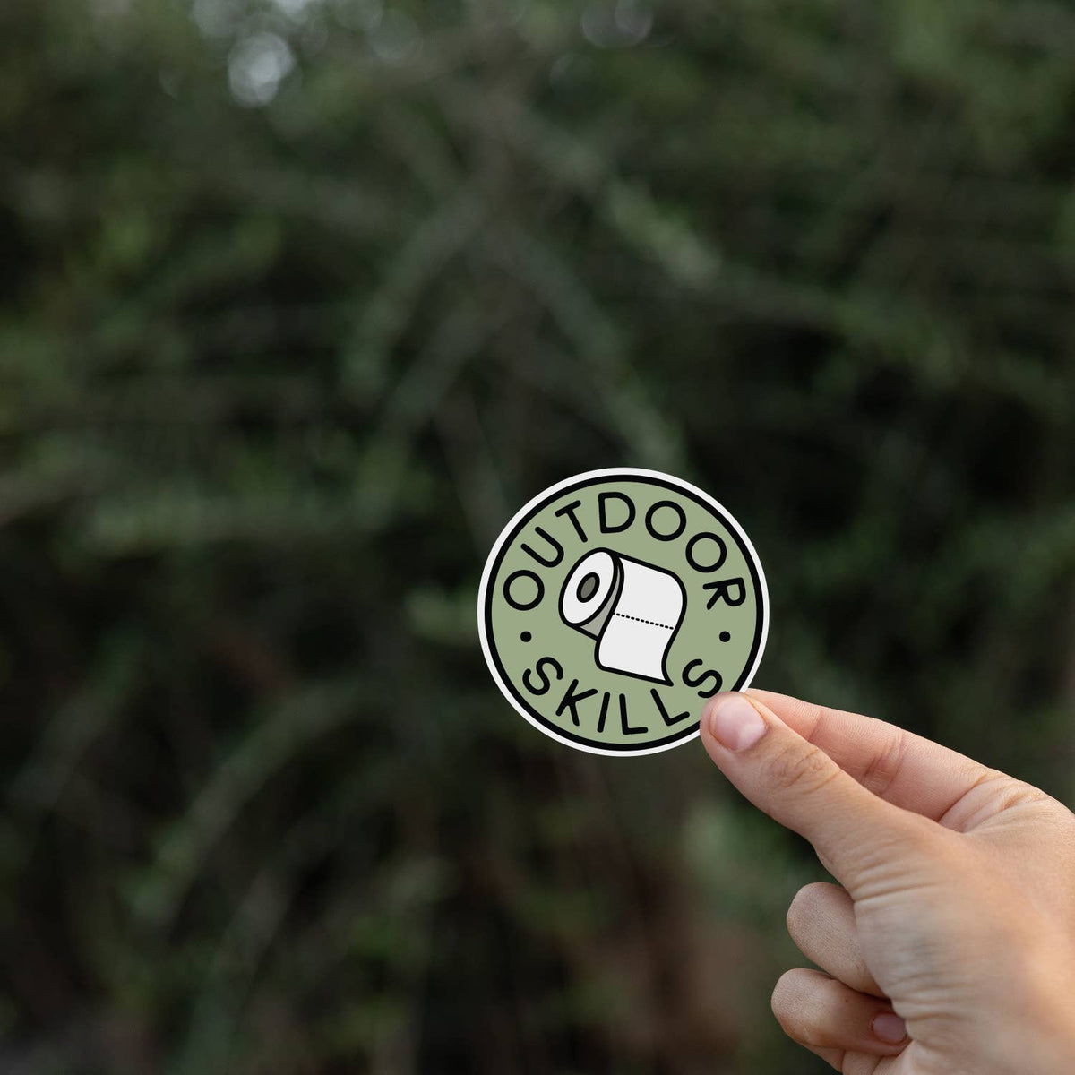 Fingers lift the Outdoor Skills sticker against a blurred wall of dark evergreen branches.
