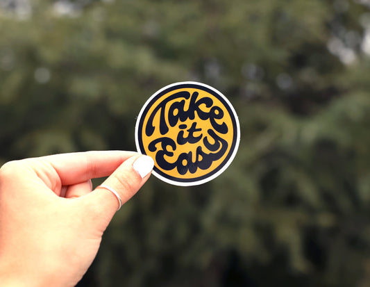 Pinched at arm's length, the Take it Easy sticker reads in black script with pine branches and sky behind.