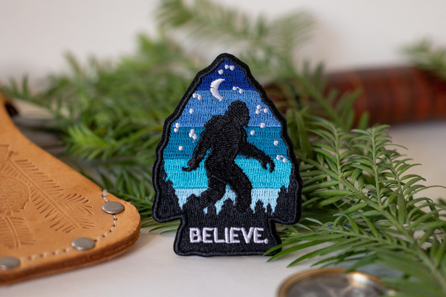 Propped among evergreen sprigs and a leather sheath, the Bigfoot Believe patch shows its moonlit blue arrowhead.