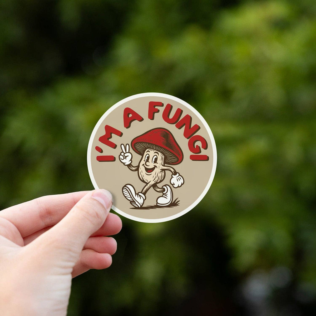 A hand holds the I'm a Fungi sticker up against blurred green leaves in soft daylight.