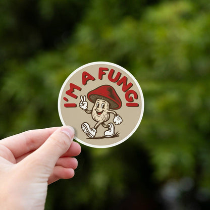A hand holds the I'm a Fungi sticker up against blurred green leaves in soft daylight.