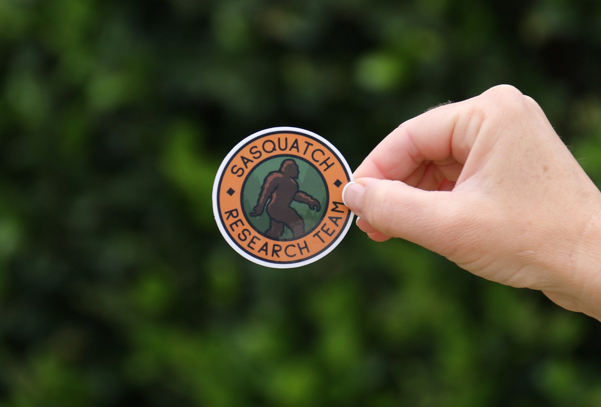 Held at arm's length, the Sasquatch Research Team sticker tilts slightly against soft green leaves.