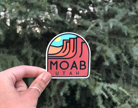 Pulled back a little, the same Moab Utah Abstract sticker sits small in the frame before a dense evergreen.