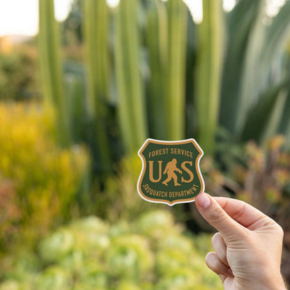 Fingers hold the US Sasquatch Department sticker before tall cactus and succulents at golden hour.
