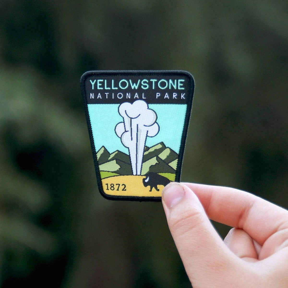 Fingers pinch the Yellowstone National Park patch in front of blurred dark evergreen branches.