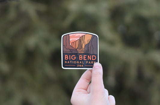Farther back against blurred evergreen branches, the Big Bend National Park sticker sits small in a hand.