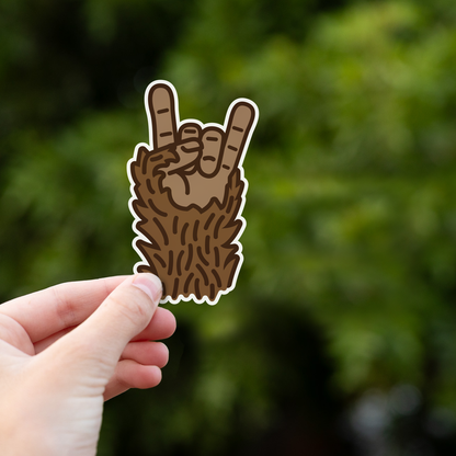 A hand holds the Bigfoot Rocks sticker up against blurred green leaves in bright daylight.