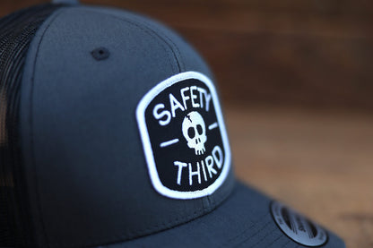 Close crop on the embroidered patch of the charcoal Safety Third trucker hat against blurred wood.