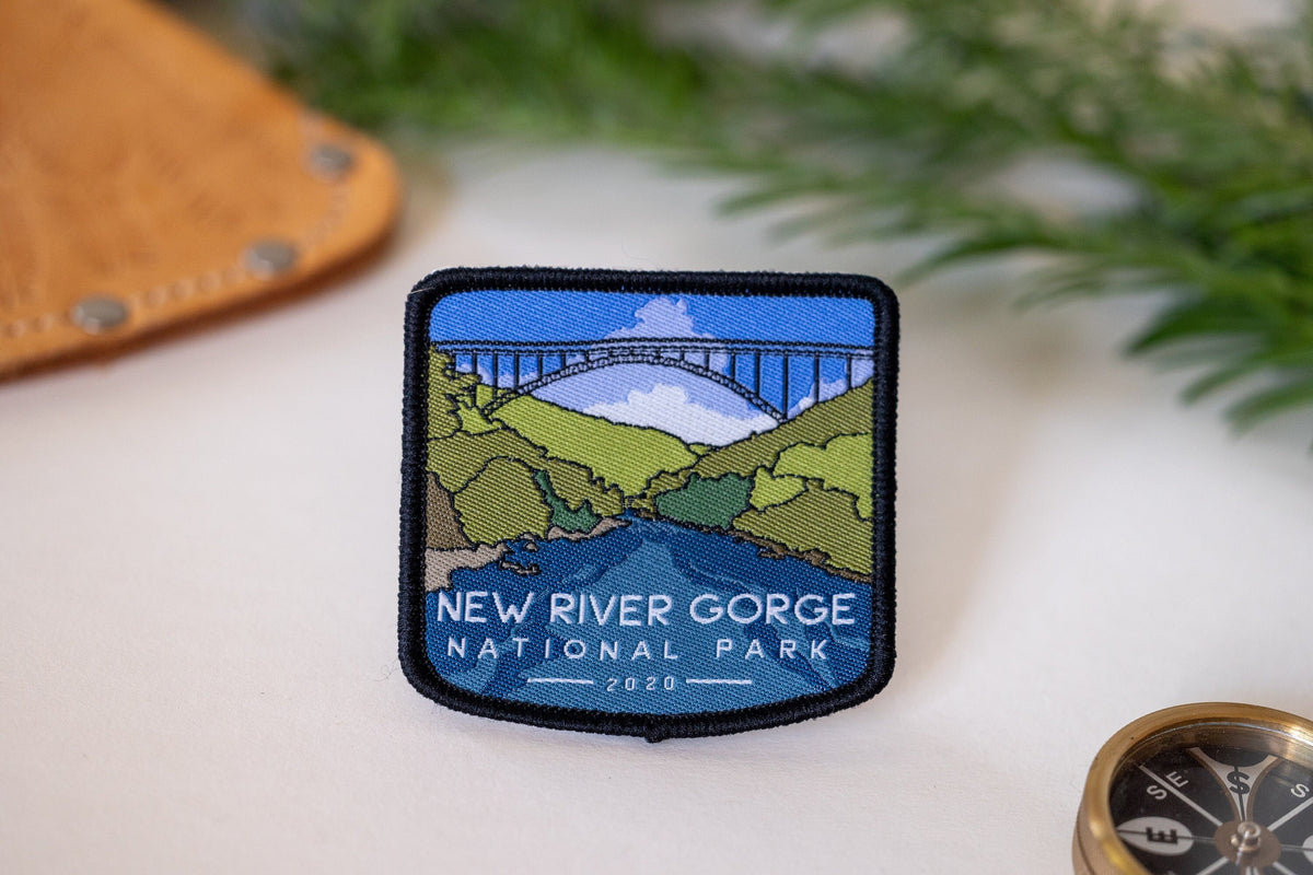 Laid on a pale surface near leather, a compass and pine sprigs, the New River Gorge National Park patch shows its bridge.