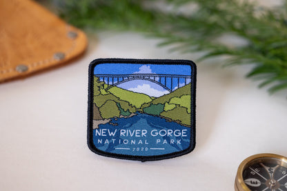 Laid on a pale surface near leather, a compass and pine sprigs, the New River Gorge National Park patch shows its bridge.