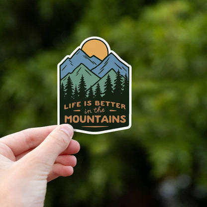 Bright green foliage sits behind the Life Is Better In The Mountains sticker pinched in a hand.