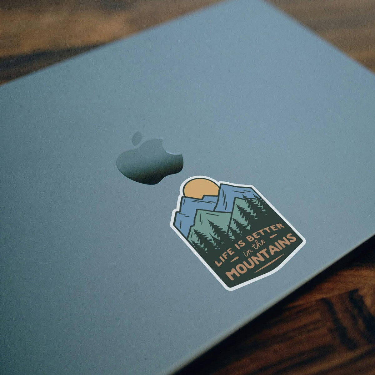 Stuck near the Apple logo on a blue laptop lid, the Life Is Better In The Mountains sticker sits at an angle.