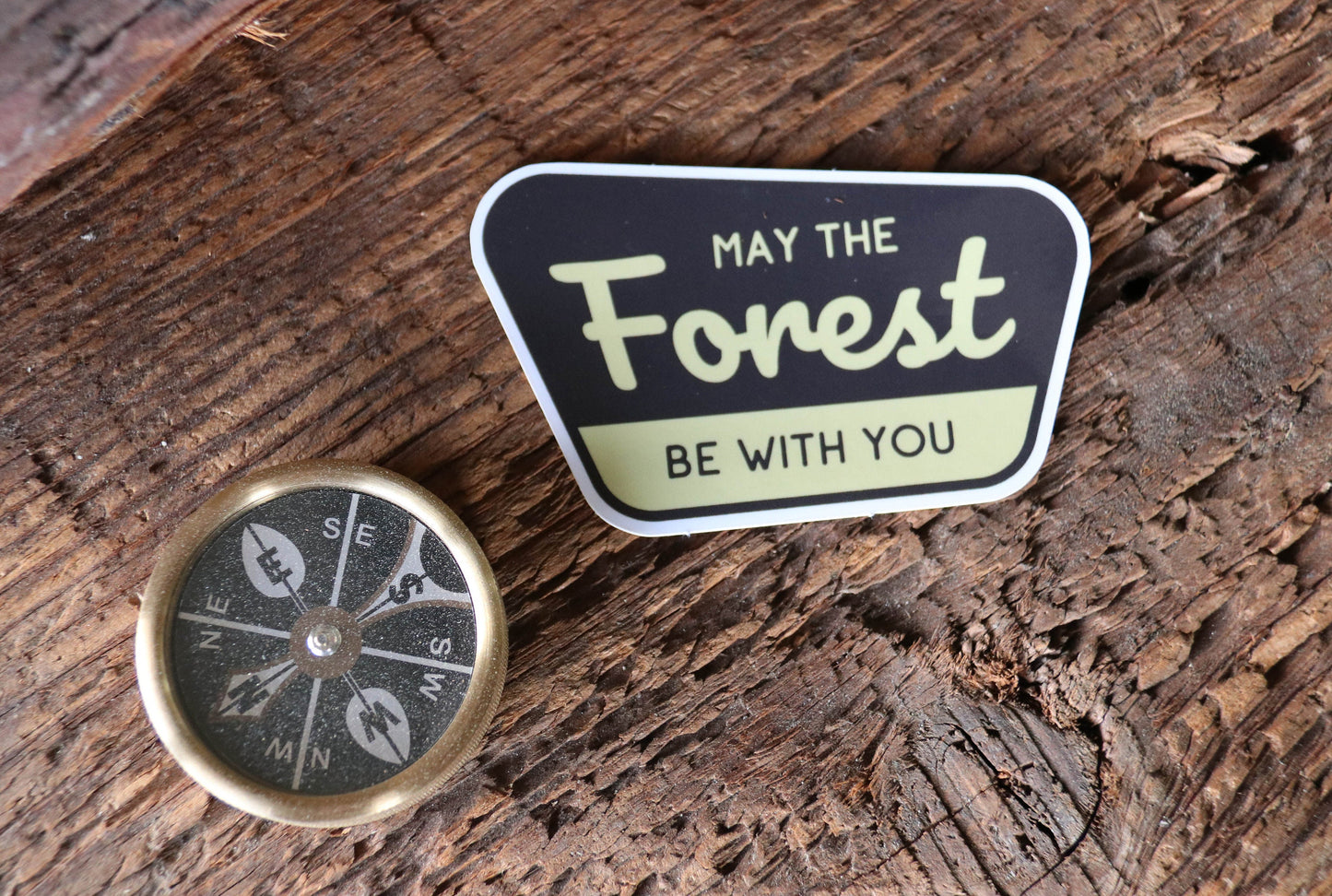 A brass compass rests on split timber beside the May The Forest Be With You sticker.