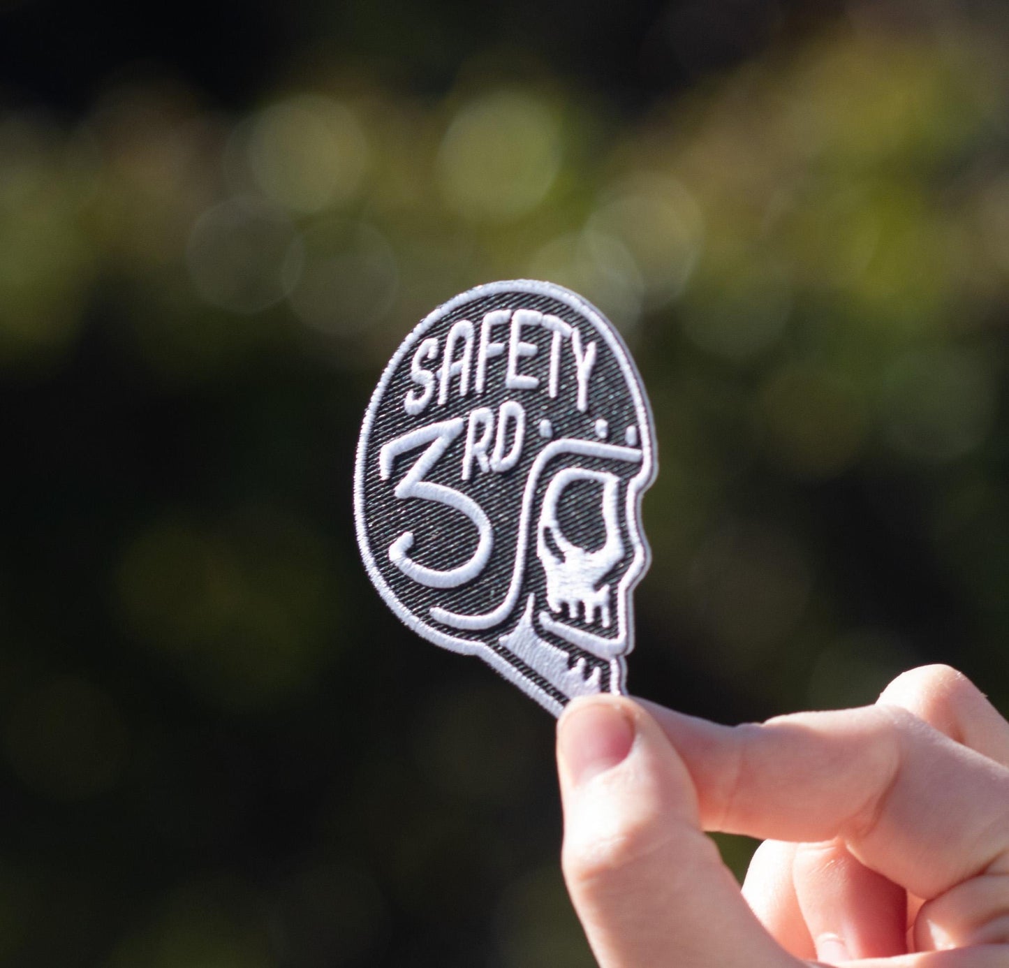 Fingertips grip the Safety 3rd Helmet Patch close to the lens, green bokeh scattered behind it.