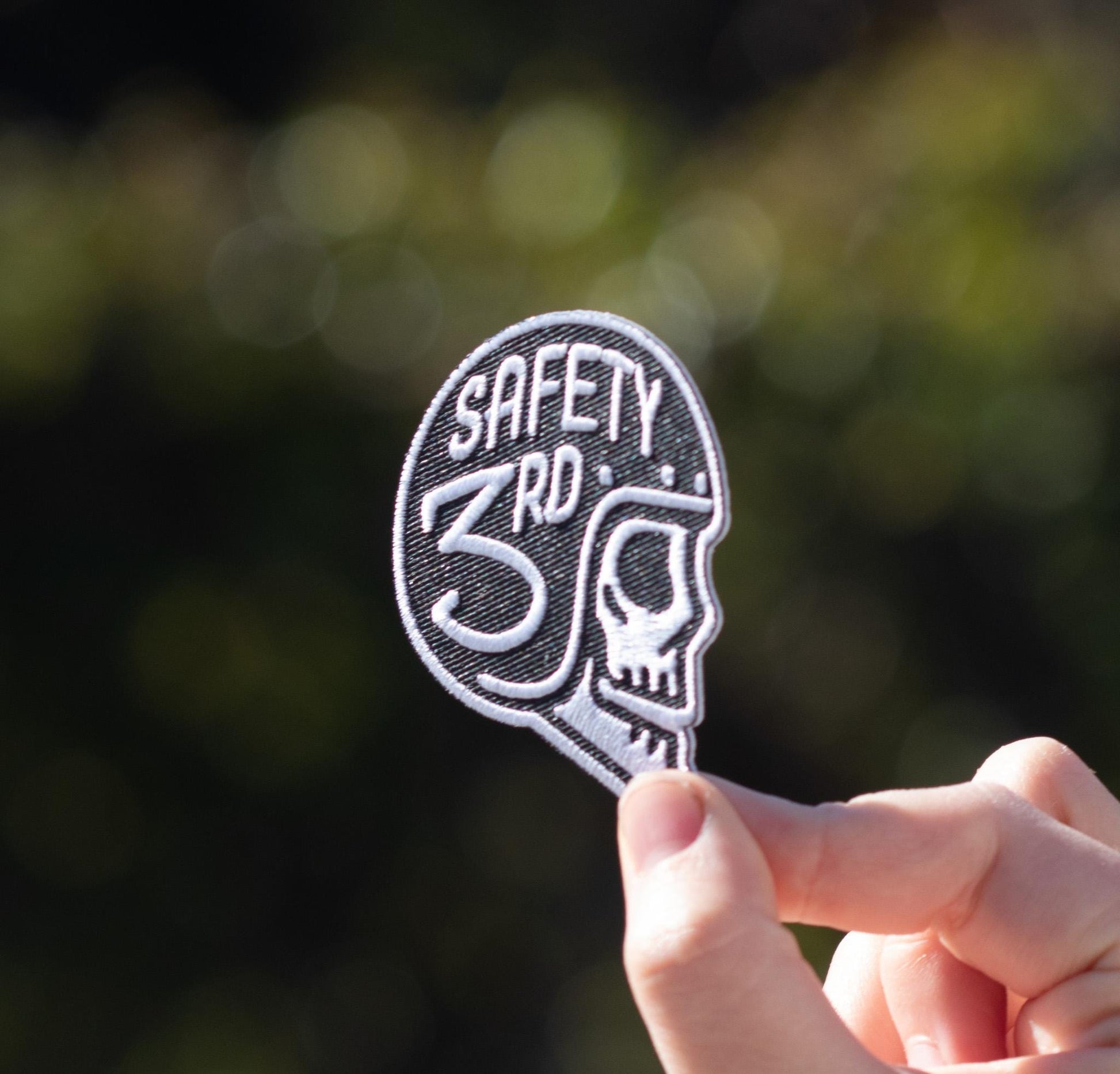 Fingertips grip the Safety 3rd Helmet Patch close to the lens, green bokeh scattered behind it.