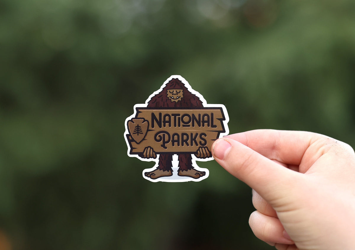 Held at arm's length, the National Parks Bigfoot sign sticker reads clearly with pine boughs far behind.