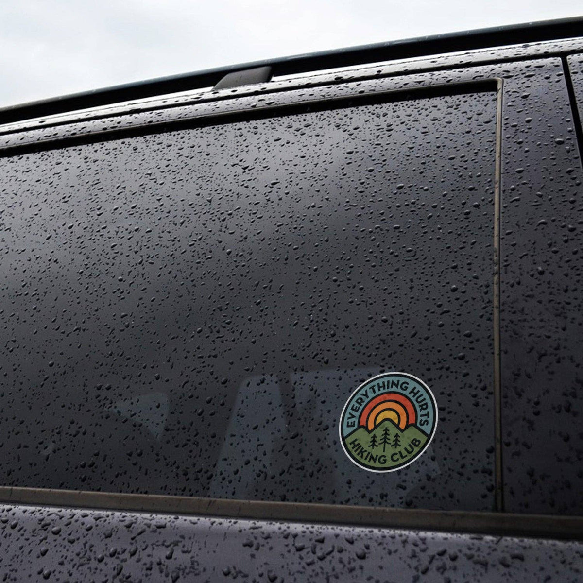 Stuck to a rain beaded car window, the Everything Hurts Hiking Club sticker sits low on the dark glass.