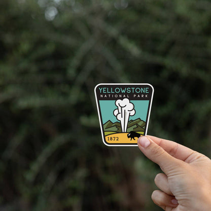 Held at arm's length toward dark evergreen branches, the Yellowstone National Park sticker catches the light.