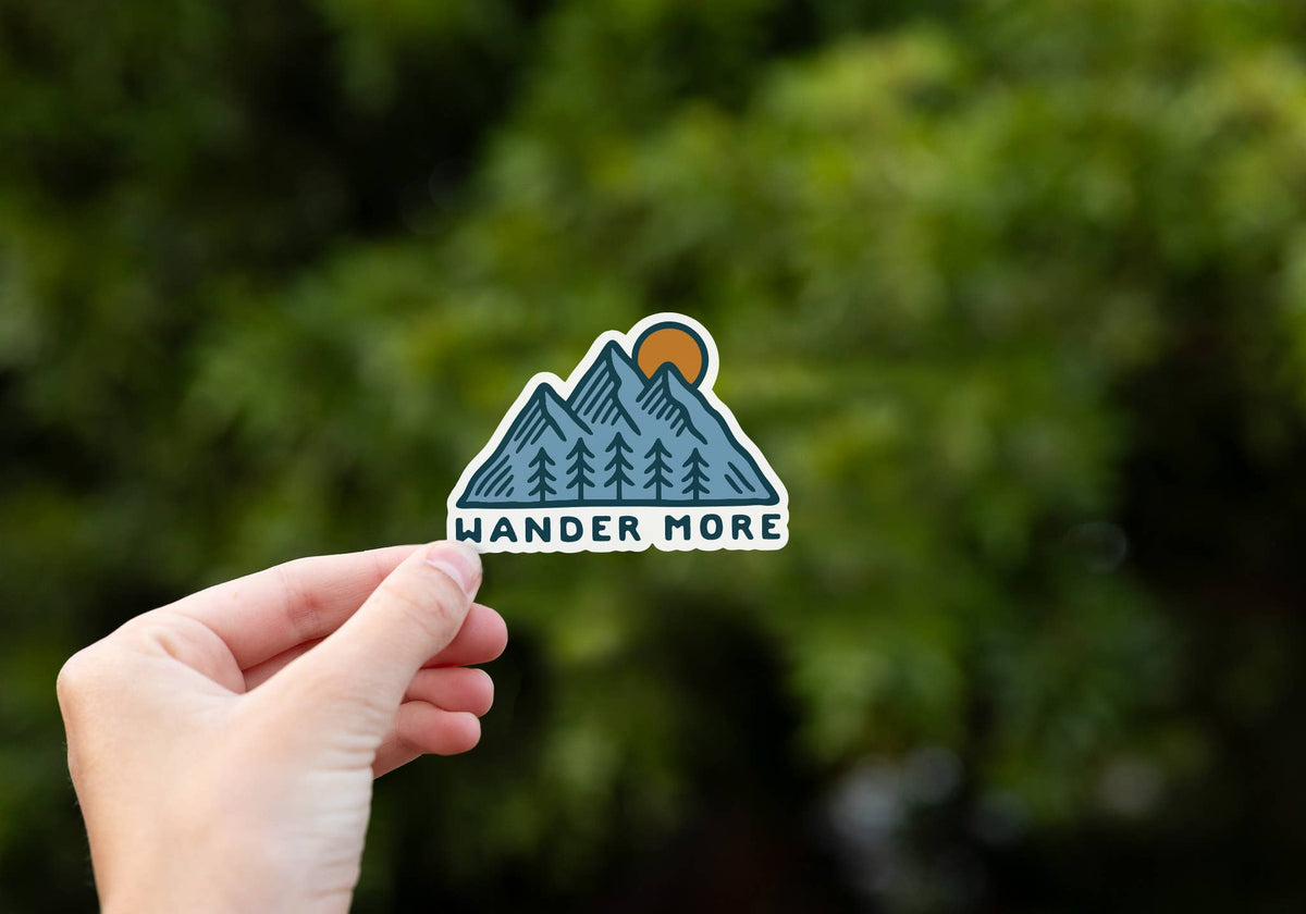 A hand holds the Wander More sticker up against blurred green trees, its mountain outline catching the light.